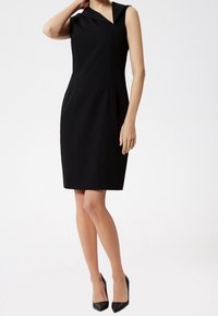 Vestido negro sin mangas con un cuello asimétrico, corte ajustado, a la altura de la rodilla y con costuras laterales sutiles. Combinado con tacones negros de punta afilada.