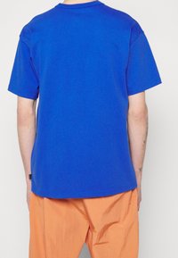 Královsky modré tričko s krátkým rukávem a kulatým výstřihem, vyrobené z bavlny, s uvolněným střihem a jednoduchým designem. Nošeno s oranžovými lehkými kalhotami.