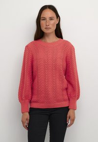 Lightweight coral knit sweater with a wave pattern, ribbed neckline and cuffs, and structured balloon sleeves. Paired with black pants.