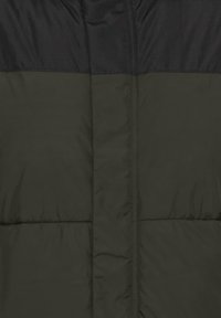 Pūšļojamā jaka ar tumši zaļu korpusu un melnu augšējo daļu. Iezīmējas ar quilted sekcijām, gludu tekstūru un ar rāvējslēdzēju aizdari. Nav redzama zīme.