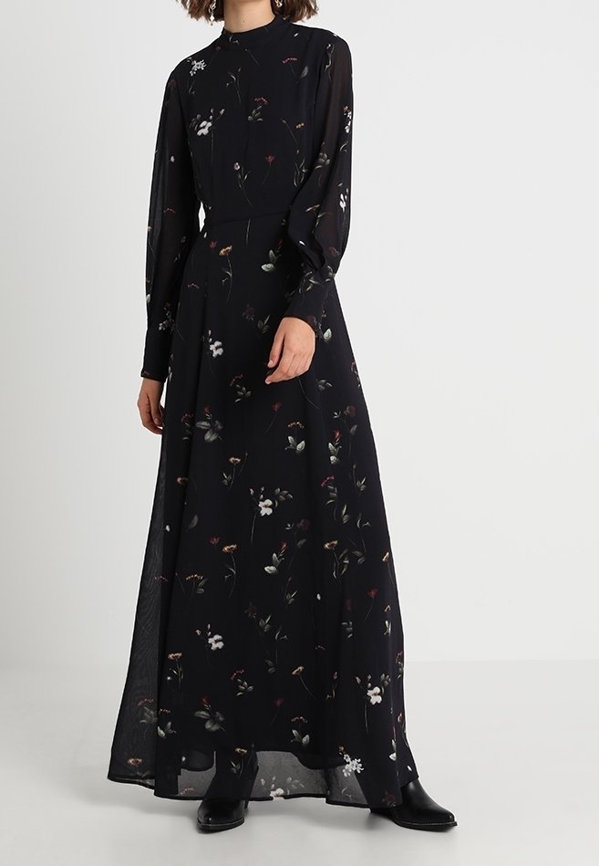 Femme portant une longue robe noire à fleurs avec des manches longues et des bottines noires, debout avec une main près de la taille.