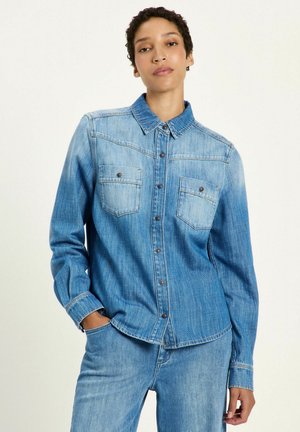 Woman wearing a faded blue denim shirt with buttoned pockets and matching denim jeans, posing with one hand in pocket against a plain background.