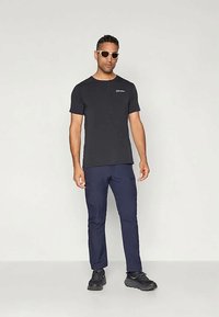 T-shirt preto de manga curta com logótipo, combinado com calças azul-marinho e ténis pretos. Os acessórios incluem óculos de sol de cor clara.