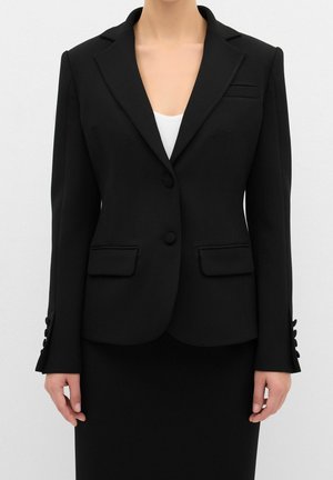 Vrouw draagt een aangesloten zwarte blazer met inkniprevers, twee klepzakken aan de voorkant en knoopmanchetten, over een wit topje en een zwarte rok.