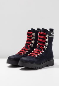 Námořnické semišové kotníkové boty s červenými tkaničkami, stříbrnými očky a texturovanou černou podrážkou. Obsahují logo na zadní straně a zesílené detaily na špičce.