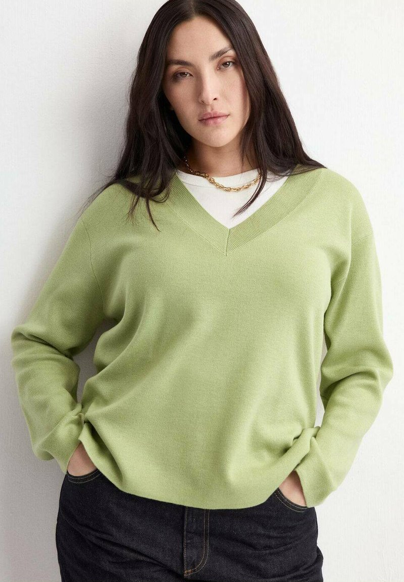 Femme aux longs cheveux noirs portant un pull vert clair à col en V par-dessus une chemise blanche et un pantalon foncé, debout les mains dans les poches contre un mur blanc.