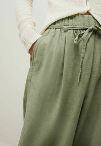 Pantaloni in lino verde chiaro con cintura elastica, lacci regolabili e tasche laterali, abbinati a un maglione crema texturizzato.