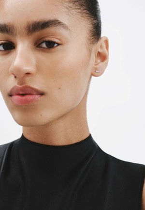 Top negro de cuello alto, primer plano de la cara; piel suave, ojos marrones, labios naturales, cabello corto y pequeño pendiente de perno. Fondo minimalista.