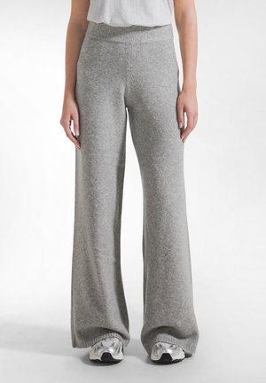 Persoon die hoog getailleerde, wijde grijze gebreide broek draagt, gecombineerd met witte en zilveren sneakers, staand tegen een effen witte achtergrond.
