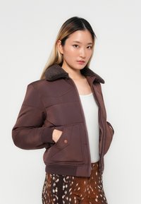 Giacca imbottita marrone con colletto in morbido pelo, chiusura a zip, polsini a costine e tasca laterale. Indossata sopra un top bianco e pantaloni a fantasia.