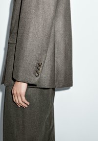 Blazer grigio su misura con spalle strutturate, quattro bottoni sul polsino e una texture liscia, abbinato a una gonna coordinata.