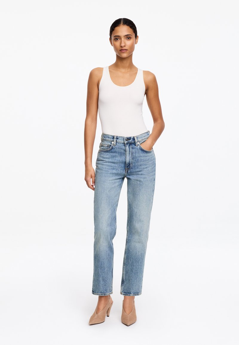 Femme debout portant un haut blanc sans manches, un jean taille haute bleu clair et des talons beiges pointus, avec une main dans la poche.