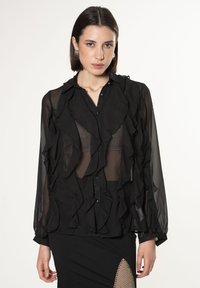 Blusa nera trasparente con dettagli a volant, maniche lunghe, chiusura a bottoni e una texture leggera. Abbinata a una gonna nera aderente.