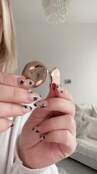 Uhani z zlatim tonom v obliki kroga z gladko, odsevno površino in zapiranjem na sponko. Opazna tekstura v kontrastu z nohti z vzorcem zvezd.