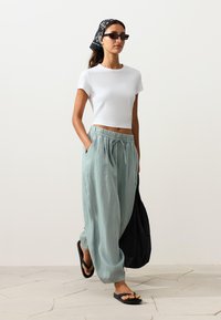 Vrouw loopt, draagt een wit cropped T-shirt, lichtblauwe losse broek, zwarte slippers, zwarte patroon hoofddoek, zonnebril en houdt een zwarte tas vast.