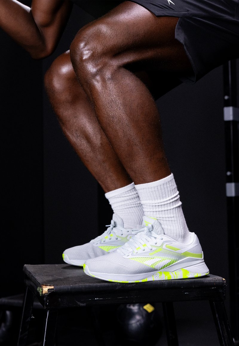 Uns sapatilhas de desporto cinzentas com detalhes amarelos, parte superior em malha e sola padronizada, acompanhadas por meias brancas, repousando numa bancada preta.