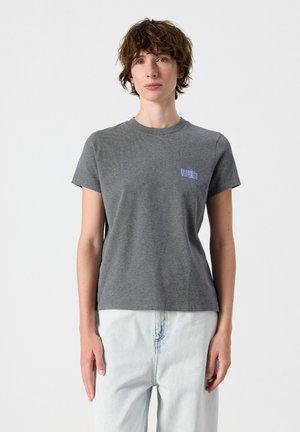 Person with short curly brown hair wearing a gray T-shirt and light blue jeans standing against a plain background.