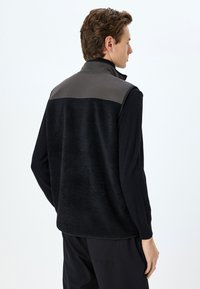 Schwarze Fleece-Weste mit grauem Schulterpanel, hohem Kragen und strukturiertem Material. Verfügt über einen Reißverschluss und elastische Bündchen. Minimalistisches Design.