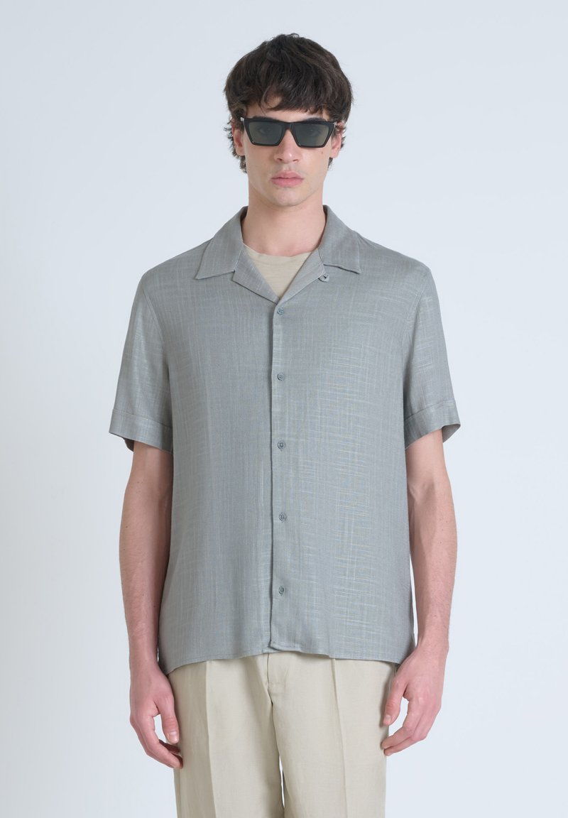 Camisa de botones de manga corta, gris claro, con tejido texturizado, cuello doblado y corte holgado, combinada con una capa inferior beige y pantalones beige.