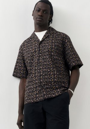 Muž s tmavou pokožkou a pletenými vlasmi, oblečený v čiernej krátkej košeli so vzorom opakujúceho sa nápisu „HUGO“ a tmavých nohaviciach, stojaci pred jednoduchým pozadím.