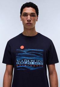 Námořnická modrá bavlněná tričko s grafickým designem vlnitých modrých linií a oranžovým sluncem. Název značky "NAPAPIJRI" je výrazně zobrazen.