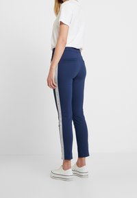 Navyblå tajta byxor med tapered passform, elastisk midja, sidofickor med dragkedja och kontrasterande sidolinje. Bärs med vita sneakers.