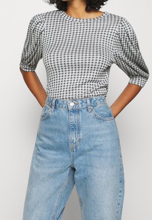 Osoba w czarno-białym topie w kratkę w stylu bufek, włożonym do wysokich jasnoniebieskich dżinsów, z rękami opartymi na biodrach.