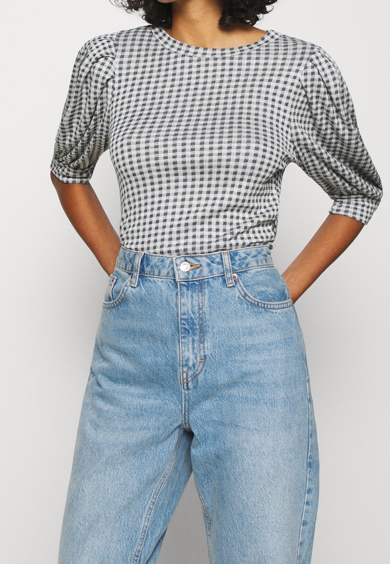 Personne portant un haut à carreaux vichy noir et blanc avec des manches bouffantes, rentré dans un jean en denim bleu clair taille haute, les mains posées sur les hanches.
