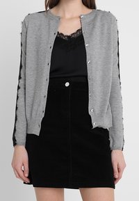 Grå cardigan med knappstängning, med texturerade svarta accentdetaljer längs ärmarna. Matchas med en svart spets camisole och svart kjol.