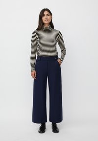 Gestreepte coltrui in zwart en wit, gecombineerd met brede marineblauwe broek met zijzakken en knoopaccenten, gestyled met zwarte schoenen.