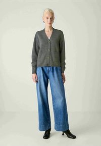 Cardigan grigio a maglia con scollatura a V, cinque bottoni e tasche anteriori, abbinato a jeans in denim blu a gamba larga e stivaletti neri.