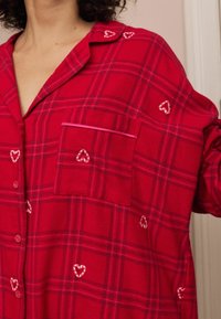 Czerwona koszula flanelowa w kratę z kieszonką, haftem w kształcie serca i kontrastowymi lamówkami. Klasyczny fason zapinany na guziki, o luźnym kroju i miękkiej fakturze.