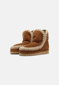Stivaletti alla caviglia in suede marrone con una texture morbida, caratterizzati da una cucitura decorativa color crema lungo il bordo superiore e una suola piatta in gomma.