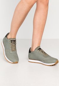 Vihreät teksturoidut lenkkarit, joissa on nauhakiinnitys, valkoinen välikerros ja kumipohja, sekä mustat yksityiskohdat kantapäässä.