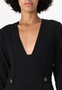 Schwarzes Kleid mit tiefem V-Ausschnitt, langen Ärmeln und plissiertem Rock. Verfügt über goldfarbene Knopfakzente in der Taille. Glatte Stofftextur.