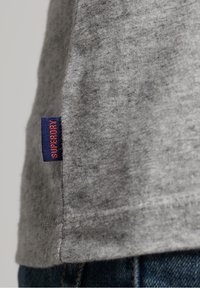 Grå tygplagg med en blå och röd etikett märkt "SUPERDRY" på sidon, bärs över blå denimjeans.