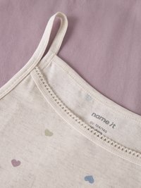 Haut beige sans manches avec de fines bretelles et de petites impressions de cœurs colorés sur un fond en tissu violet.