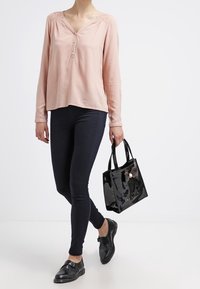 Vaaleanpunainen pitkähihaista puseroa, jossa on V-pääntie ja napitus, yhdistetty tummiin slim fit -farkkuihin, mustiin kenkiin ja pieneen mustaan kiiltävään käsilaukkuun.