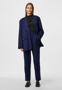 Jūras spēku blazeris un pielāgoti bikses gludā audumā. Blazerim ir brīvs piegriezums ar lieliem atlokiem un kabatām, ko papildina melnas kurpes.