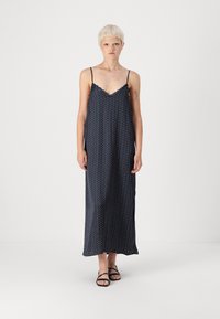 Navy blue sleeveless maxi dress with polka dots, lace trim neckline, and flowing fabric. Features thin adjustable straps and side slits.