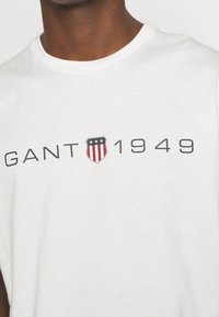 Man wearing white T-shirt with "GANT 1949" text and shield logo with stars and stripes in red, white, and blue on the chest.