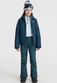 Tenue de ski comprenant une veste isolante bleu foncé, une couche de base zippée bleu clair, un pantalon assorti, des lunettes de ski noires et un bonnet avec pompon bleu.