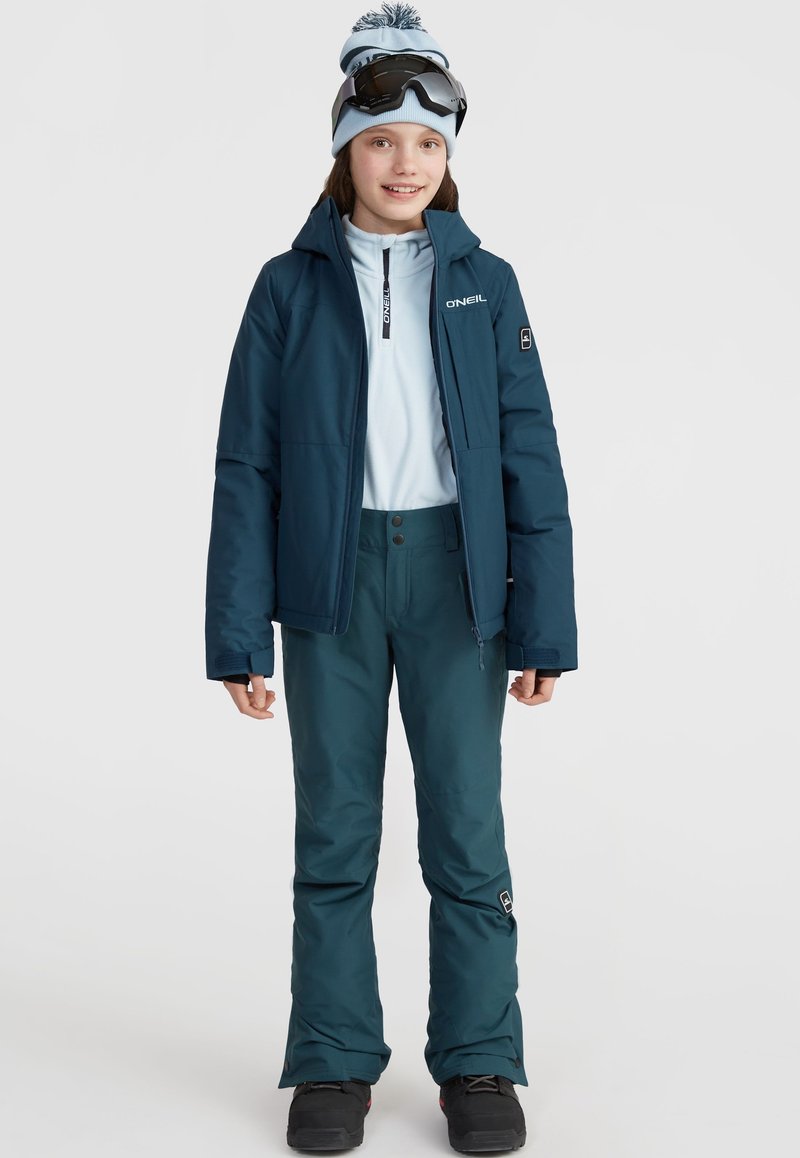 Tenue de ski comprenant une veste isolante bleu foncé, une couche de base zippée bleu clair, un pantalon assorti, des lunettes de ski noires et un bonnet avec pompon bleu.
