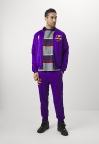 Conjunto desportivo roxo com logótipo amarelo "Lakers" na jaqueta e nas calças. Camiseta cinza listrada por baixo. Ténis pretos completam o visual.