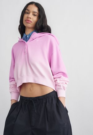 Kobieta o długich ciemnych włosach nosi różową krótką bluzę z kapturem na niebieskiej koszuli i luźne czarne spodnie, trzymając ręce w kieszeniach na jednolitym tle.