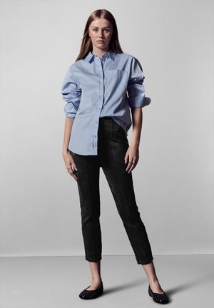 Chemise légère bleue à manches retroussées, associée à un pantalon cropped en daim noir et des chaussures plates noires ornées de clous.
