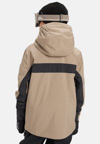 Osoba oblečená v béžovo-čiernej kárované lyžiarskej bunde, čiernej pletenej čiapke, čiernych rukaviciach a béžových lyžiarskych okuliaroch, zachytená zozadu.