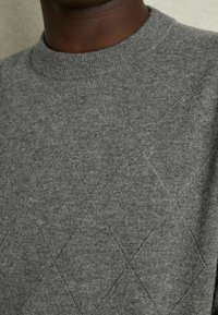 Pull en tricot gris avec un motif en losanges, un col côtelé et un tissu texturé. Gros plan sur le matériau et les détails de couture.