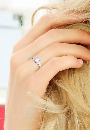 Bague en argent avec une pierre précieuse ronde et claire, bande lisse avec une légère courbure, exposée sur une main reposant contre des cheveux de couleur claire.