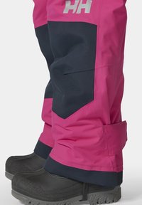 Růžové a námořnické voděodolné sněhové kalhoty mají volný střih, zesílené manžety a texturované panely s detailem loga. Černé sněhové boty s robustními podrážkami.